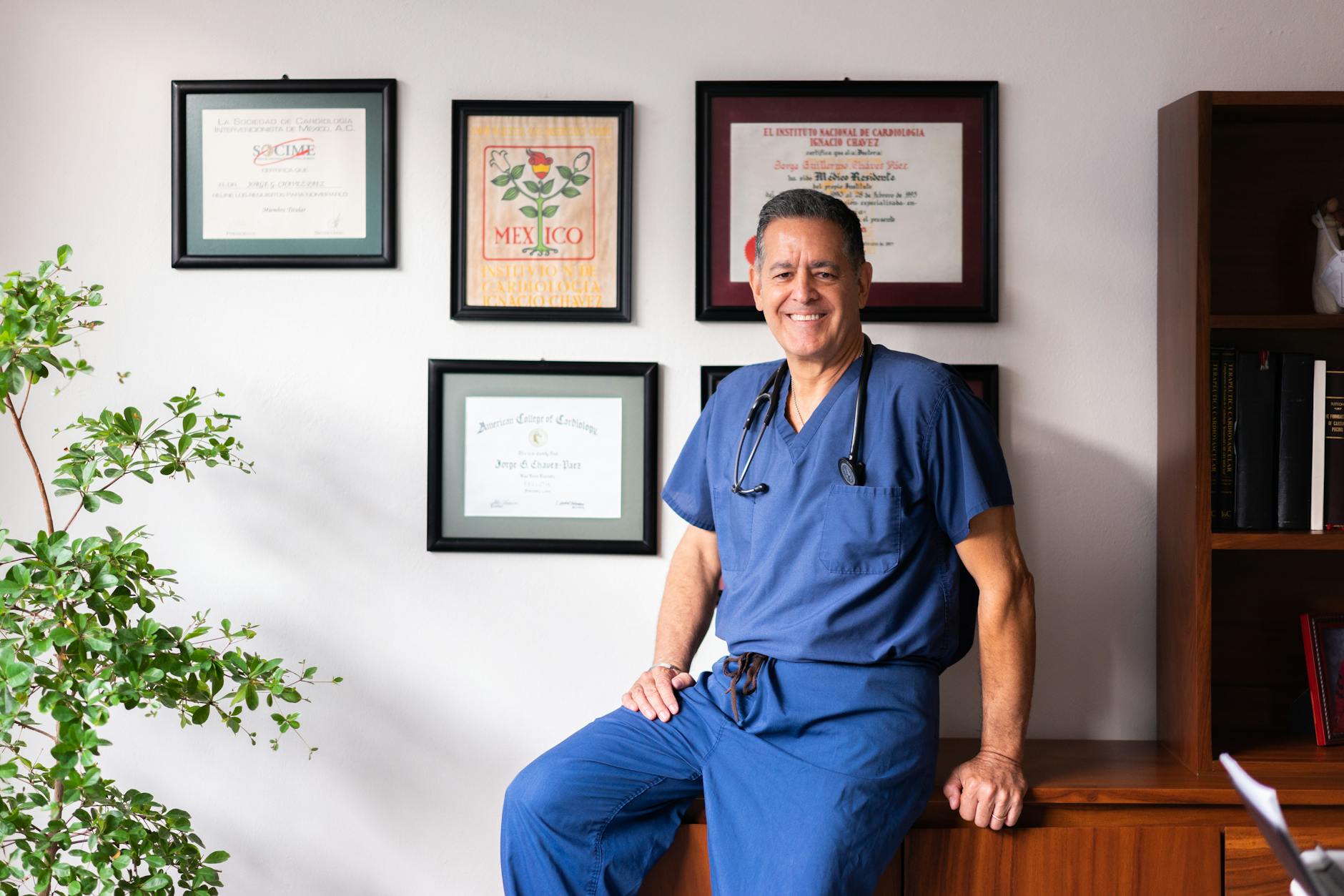 A smiling doctor in scrubs poses in his clinic, surrounded by framed certifications.